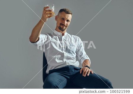 Man with stylish mustache, in classic white shirt and blue trousers is sitting on chair, holding glass of whiskey. Grey background, close-up shot. Man with stylish mustache, in classic white shirt and blue trousers is sitting on chair, holding glass of whiskey. Grey background, close-up shot. 100321599