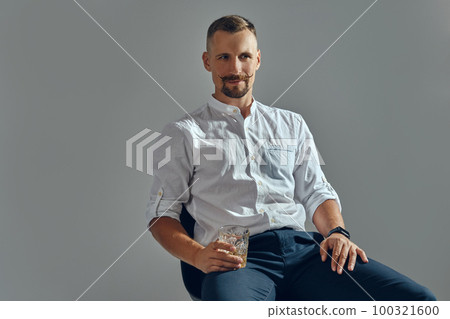 Man with stylish mustache, in classic white shirt and blue trousers is sitting on chair, holding glass of whiskey. Grey background, close-up shot. Man with stylish mustache, in classic white shirt and blue trousers is sitting on chair, holding glass of whiskey. Grey background, close-up shot. 100321600