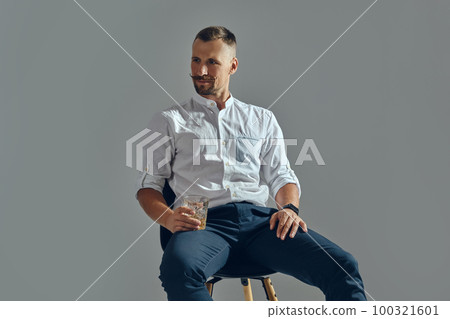Man with stylish mustache, in classic white shirt and blue trousers is sitting on chair, holding glass of whiskey. Grey background, close-up shot. Man with stylish mustache, in classic white shirt and blue trousers is sitting on chair, holding glass of whiskey. Grey background, close-up shot. 100321601