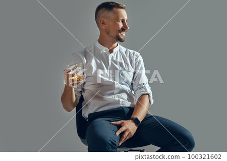Man with stylish mustache, in classic white shirt and blue trousers is sitting on chair, holding glass of whiskey. Grey background, close-up shot. 100321602