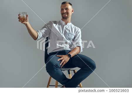 Man with stylish mustache, in classic white shirt and blue trousers is sitting on chair, holding glass of whiskey. Grey background, close-up shot. 100321603