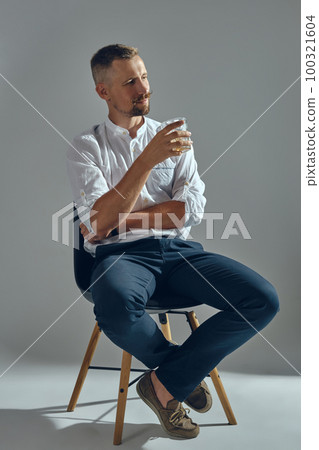 Man with stylish mustache, in classic white shirt and blue trousers is sitting on chair, holding glass of whiskey. Grey background, close-up shot. Man with stylish mustache, in classic white shirt and blue trousers is sitting on chair, holding glass of whiskey. Grey background, close-up shot. 100321604
