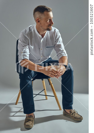 Man with stylish mustache, in classic white shirt and blue trousers is sitting on chair, holding glass of whiskey. Grey background, close-up shot. Man with stylish mustache, in classic white shirt and blue trousers is sitting on chair, holding glass of whiskey. Grey background, close-up shot. 100321607