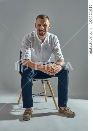 Man with stylish mustache, in classic white shirt and blue trousers is sitting on chair, holding glass of whiskey. Grey background, close-up shot. Man with stylish mustache, in classic white shirt and blue trousers is sitting on chair, holding glass of whiskey. Grey background, close-up shot. 100321612