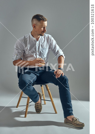 Man with stylish mustache, in classic white shirt and blue trousers is sitting on chair, holding glass of whiskey. Grey background, close-up shot. Man with stylish mustache, in classic white shirt and blue trousers is sitting on chair, holding glass of whiskey. Grey background, close-up shot. 100321618