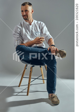 Man with stylish mustache, in classic white shirt and blue trousers is sitting on chair, holding glass of whiskey. Grey background, close-up shot. Man with stylish mustache, in classic white shirt and blue trousers is sitting on chair, holding glass of whiskey. Grey background, close-up shot. 100321620