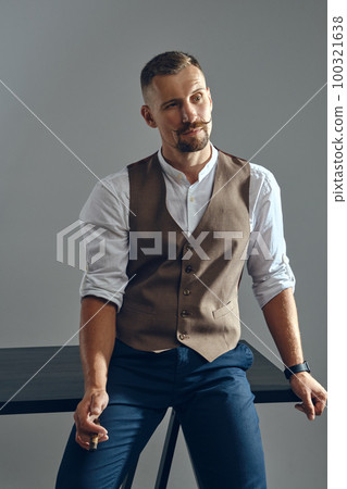 Man with a stylish mustache, dressed in brown vest, white shirt and dark trousers is sitting on table, enjoying cigar. Grey background, close-up. Man with a stylish mustache, dressed in brown vest, white shirt and dark trousers is sitting on table, enjoying cigar. Grey background, close-up. 100321638