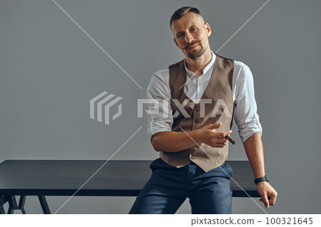 Man with a stylish mustache, dressed in brown vest, white shirt and dark trousers is sitting on table, enjoying cigar. Grey background, close-up. Man with a stylish mustache, dressed in brown vest, white shirt and dark trousers is sitting on table, enjoying cigar. Grey background, close-up. 100321645