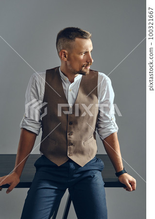 Man with a stylish mustache, dressed in brown vest, white shirt and dark trousers is posing sitting on table. Grey background, close-up. Man with a stylish mustache, dressed in brown vest, white shirt and dark trousers is posing sitting on table. Grey background, close-up. 100321667