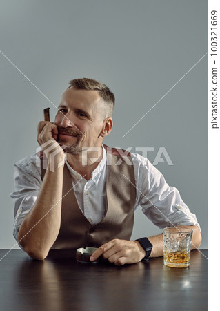 Man with stylish mustache, dressed in classic brown vest, white shirt is sitting at table, enjoying whiskey, cigar. Grey background, close-up shot. 100321669