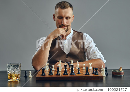 Man with stylish mustache, dressed in brown vest, white shirt is playing chess. Nearby glass of whiskey, ashtray, cigar. Grey background, close-up. Man with stylish mustache, dressed in brown vest, white shirt is playing chess. Nearby glass of whiskey, ashtray, cigar. Grey background, close-up. 100321727