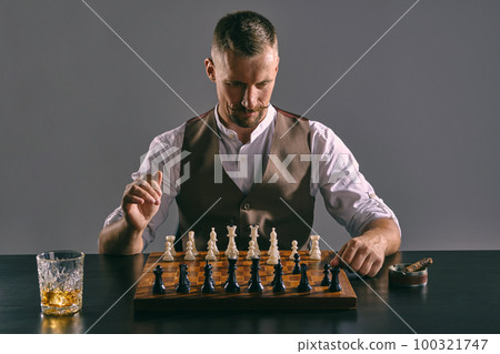 Man with stylish mustache, dressed in brown vest, white shirt is playing chess. Nearby glass of whiskey, ashtray, cigar. Grey background, close-up. Man with stylish mustache, dressed in brown vest, white shirt is playing chess. Nearby glass of whiskey, ashtray, cigar. Grey background, close-up. 100321747