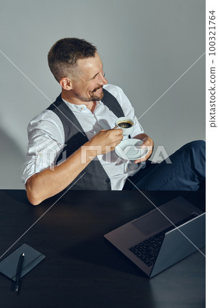 Businessman with stylish mustache, dressed in classic suit is smiling, looking away while sitting at table in office and having coffee. Top view. 100321764