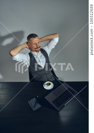 Bearded businessman with stylish mustache, dressed in classic suit is getting relax while sitting at a table in office, gray background. Top view. Bearded businessman with stylish mustache, dressed in classic suit is getting relax while sitting at a table in office, gray background. Top view. 100322689