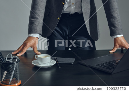 Close-up shot of businessman hands, he is posing standing at the table with a laptop on it, in office, gray background. Dressed in a classic suit. 100322750