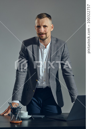 Bearded businessman with stylish mustache, dressed in a classic suit is working at laptop while sitting at table in office, gray background. 100322757