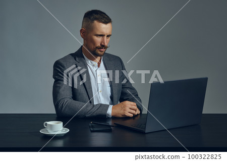 Bearded businessman with stylish mustache, dressed in a classic suit is working at laptop while sitting at table in office, gray background. Bearded businessman with stylish mustache, dressed in a classic suit is working at laptop while sitting at table in office, gray background. 100322825