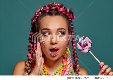 Lovely girl with a multi-colored braids hairstyle and bright make-up, posing in studio against a blue background, holding a lollipop in her hand. Lovely girl with a multi-colored braids hairstyle and bright make-up, posing in studio against a blue background, holding a lollipop in her hand. 100322942