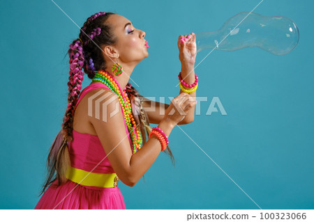 Lovely girl with a multi-colored braids hairstyle and bright make-up, is blowing bubbles using her hands, posing in studio against a blue background. 100323066
