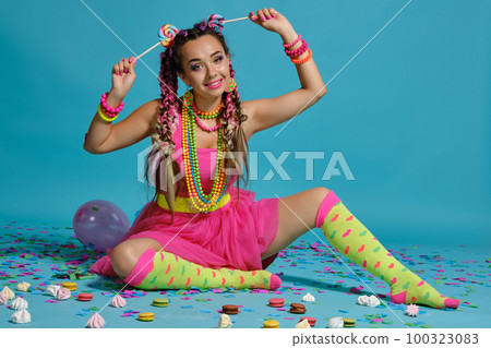 Lovely girl with a multi-colored braids hairstyle and bright make-up, posing in studio with lollipop, air balloons and confetti against a blue background. 100323083