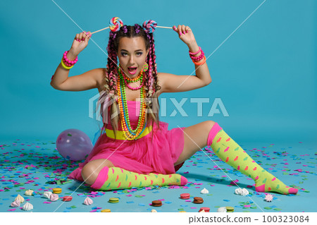 Lovely girl with a multi-colored braids hairstyle and bright make-up, posing in studio with lollipop, air balloons and confetti against a blue background. Lovely girl with a multi-colored braids hairstyle and bright make-up, posing in studio with lollipop, air balloons and confetti against a blue background. 100323084