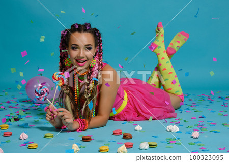 Lovely girl with a multi-colored braids hairstyle and bright make-up, posing in studio with lollipop, air balloons and confetti against a blue background. Lovely girl with a multi-colored braids hairstyle and bright make-up, posing in studio with lollipop, air balloons and confetti against a blue background. 100323095