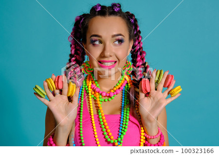 Lovely girl with multi-colored braids hairstyle and bright make-up, holding macarons between her fingers, posing in studio against a blue background. Lovely girl with multi-colored braids hairstyle and bright make-up, holding macarons between her fingers, posing in studio against a blue background. 100323166