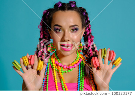 Lovely girl with multi-colored braids hairstyle and bright make-up, holding macarons between her fingers, posing in studio against a blue background. 100323177