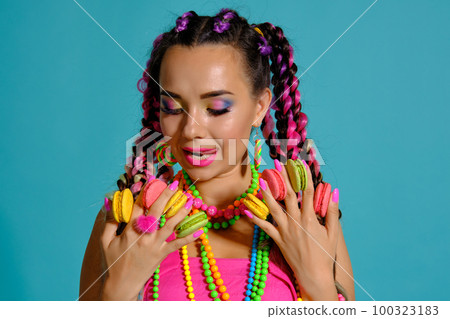Lovely girl with multi-colored braids hairstyle and bright make-up, holding macarons between her fingers, posing in studio against a blue background. 100323183