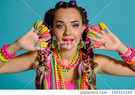 Lovely girl with multi-colored braids hairstyle and bright make-up, holding macarons between her fingers, posing in studio against a blue background. 100323184