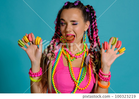 Lovely girl with multi-colored braids hairstyle and bright make-up, holding macarons between her fingers, posing in studio against a blue background. 100323219