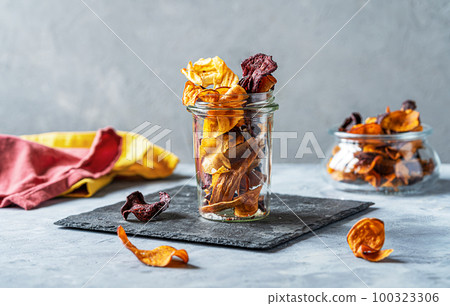Healthy vegetable chips in a glass jar on stone cutting board. Concrete background Healthy vegetable chips in a glass jar on stone cutting board. Concrete background 100323306