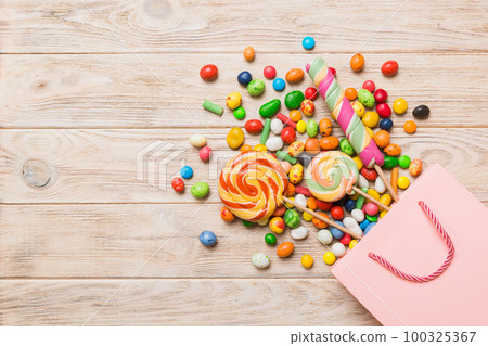 shopping paper gift bag in corner full of assorted traditional candies falling out on colored background with copy space. Happy Holidays sale concept shopping paper gift bag in corner full of assorted traditional candies falling out on colored background with copy space. Happy Holidays sale concept 100325367