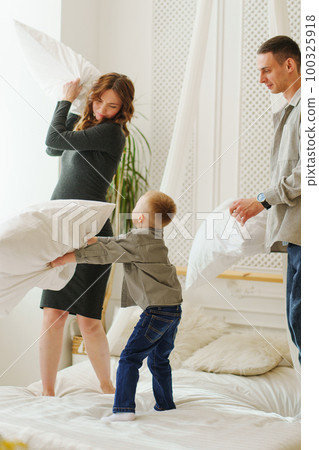 Happy pregnant mom, dad and son playing in the bedroom, fighting with pillows. Happy pregnant mom, dad and son playing in the bedroom, fighting with pillows. 100325918