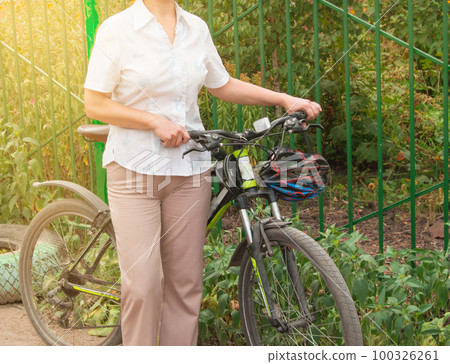Middle-aged attractive slim woman in light trousers and shirt stands near the bike in the Park on a Sunny summer day, Cycling concept for adult women 100326261
