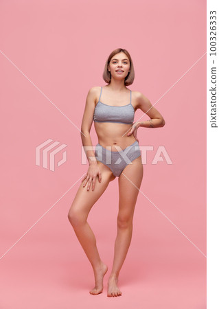 Portrait of young thin sportive woman in grey cotton inner wear posing over pink background. Concept of natural beauty, body and skin care, healthy eating Portrait of young thin sportive woman in grey cotton inner wear posing over pink background. Concept of natural beauty, body and skin care, healthy eating 100326333