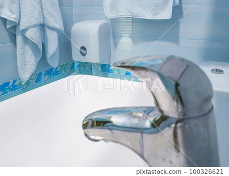 In the foreground, there is a chrome faucet in focus, a bathroom with a toilet paper dispenser, towels on the wall and a white sink. Blue tinted background In the foreground, there is a chrome faucet in focus, a bathroom with a toilet paper dispenser, towels on the wall and a white sink. Blue tinted background 100326621