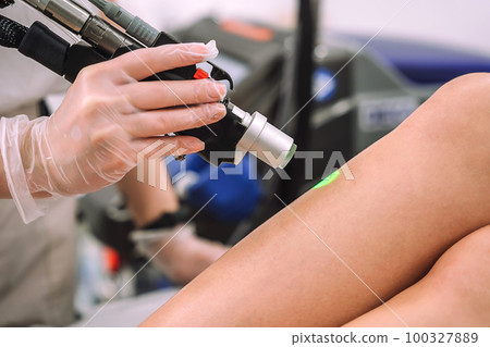 Close up shot of gloved hand with the laser hair removal machine's handpiece along woman leg. Alexandrite laser techhnology removing hair. Beauty procedure Close up shot of gloved hand with the laser hair removal machine's handpiece along woman leg. Alexandrite laser techhnology removing hair. Beauty procedure 100327889