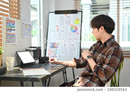 Young asian male web developer in plaid shirt using digital tablet and working with layout app design project at office 100329563