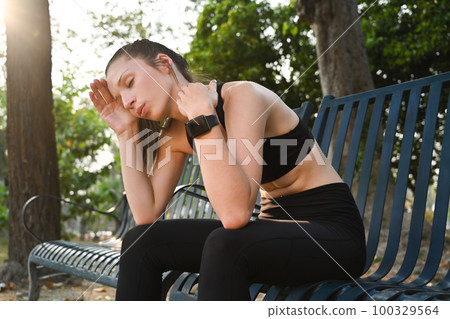 Tired athletic woman wiping sweat, resting on bench in city park after intensive fitness exercise 100329564