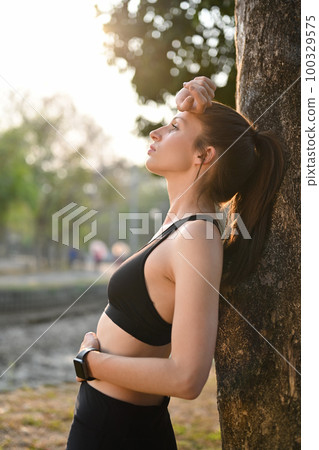 Tired fitness woman resting outdoors in park and leaning on tree trunk. Healthy lifestyle and sport concept 100329575