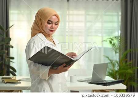 Portrait of successful muslim businesswoman standing in her office and reading information on document 100329579