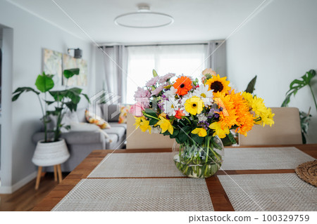 Kitchen counter table with focus on vase with huge multicolor various flower bouquet with blurred background of modern cozy living room with couch and green plants. Open space home interior design. 100329759