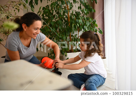 Beautiful African American woman loving mother teaching her adorable daughter to care for houseplants, smiling, enjoying together gardening in spring in a home veranda. People and environment concept 100329815