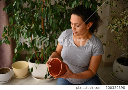 Multi-ethnic pretty woman, amateur gardener floriculturist horticulturist holding clay pot, enjoying floricultural hobby indoor, repotting houseplants at springtime. Environmental conservation concept Multi-ethnic pretty woman, amateur gardener floriculturist horticulturist holding clay pot, enjoying floricultural hobby indoor, repotting houseplants at springtime. Environmental conservation concept 100329898
