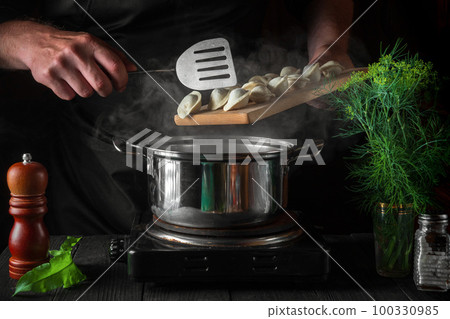 The chef cooks dumplings in a saucepan in the restaurant kitchen. Close-up of the hands of the cook during work The chef cooks dumplings in a saucepan in the restaurant kitchen. Close-up of the hands of the cook during work 100330985