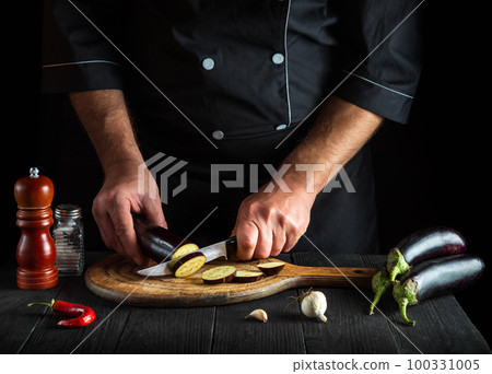 Cook cutting an eggplant in restaurant kitchen. Close-up of the hands of the chef during work. Delicious breakfast or dinner 100331005