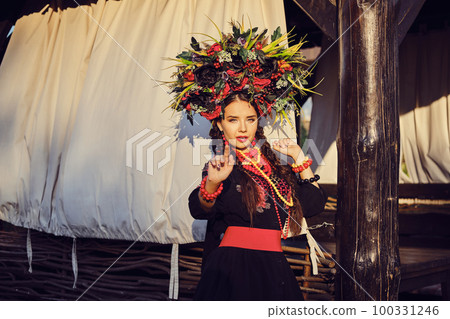 Brunette girl in a white ukrainian authentic national costume and a wreath of flowers is posing against a terrace. Brunette girl in a white ukrainian authentic national costume and a wreath of flowers is posing against a terrace. 100331246