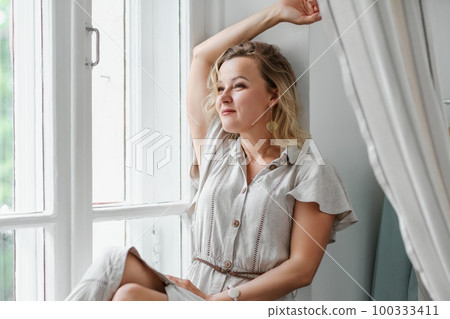 A middle-aged woman in a cream dress sits mysteriously and looks out the window on the windowsill. Green trees outside. A middle-aged woman in a cream dress sits mysteriously and looks out the window on the windowsill. Green trees outside. 100333411