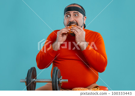 Middle-aged man with beard, dressed in a red turtleneck, headband, posing with burgers and french fries. Blue background. Close-up. Fast food. 100333443